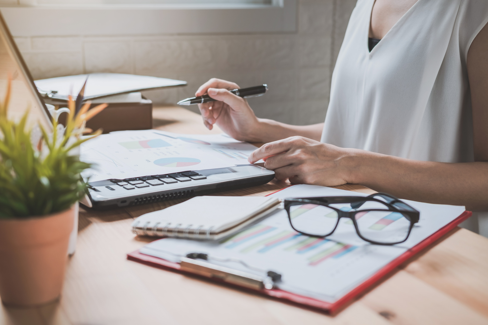 A person working at a desk with charts, a calculator, notebook, glasses, and a potted plant, demonstrating how to successfully work from home while analyzing financial documents.