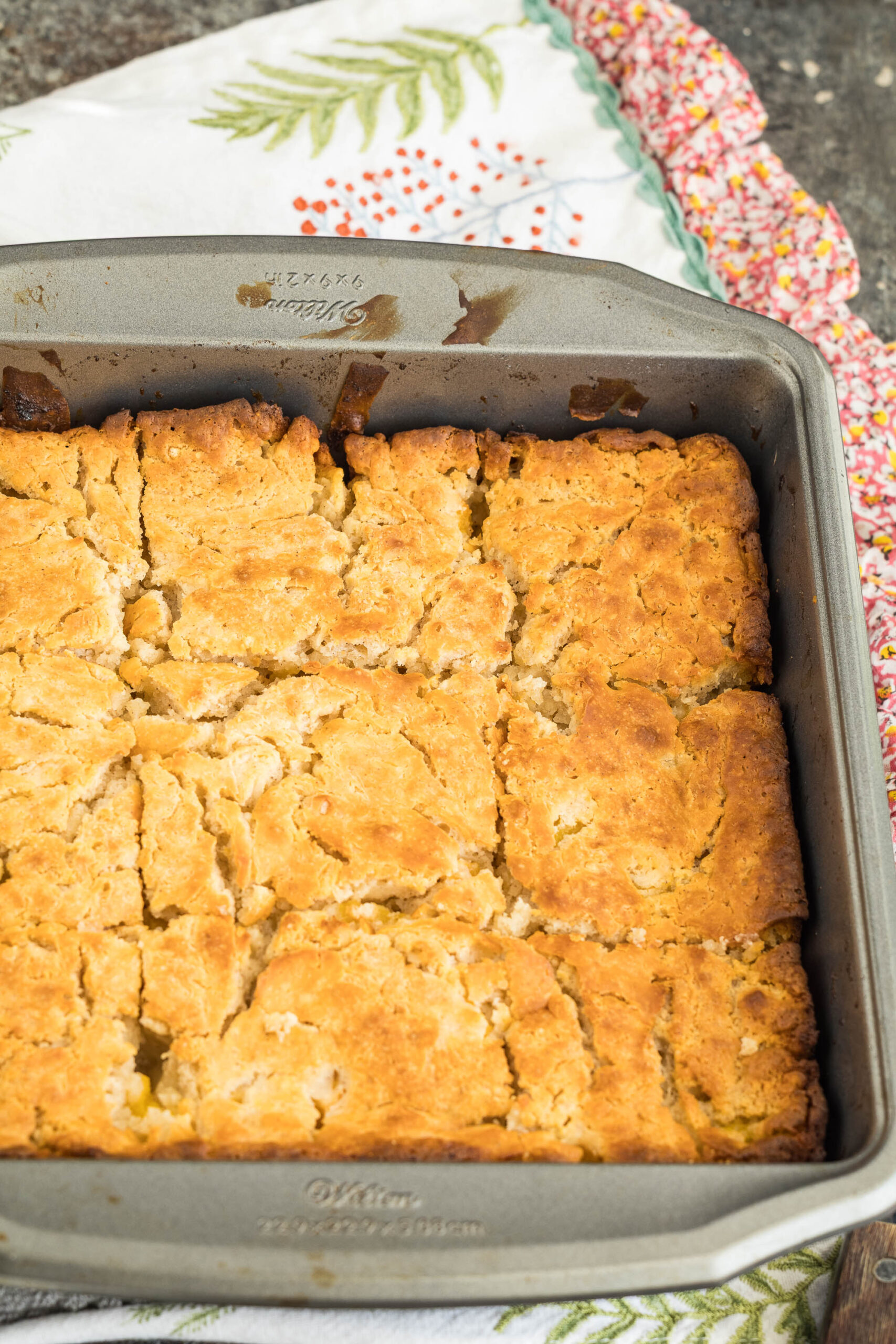 A golden-brown cobbler, reminiscent of Peach Butter Swim Biscuits, is cut into squares and sits in a metal baking dish on top of colorful and patterned kitchen towels.