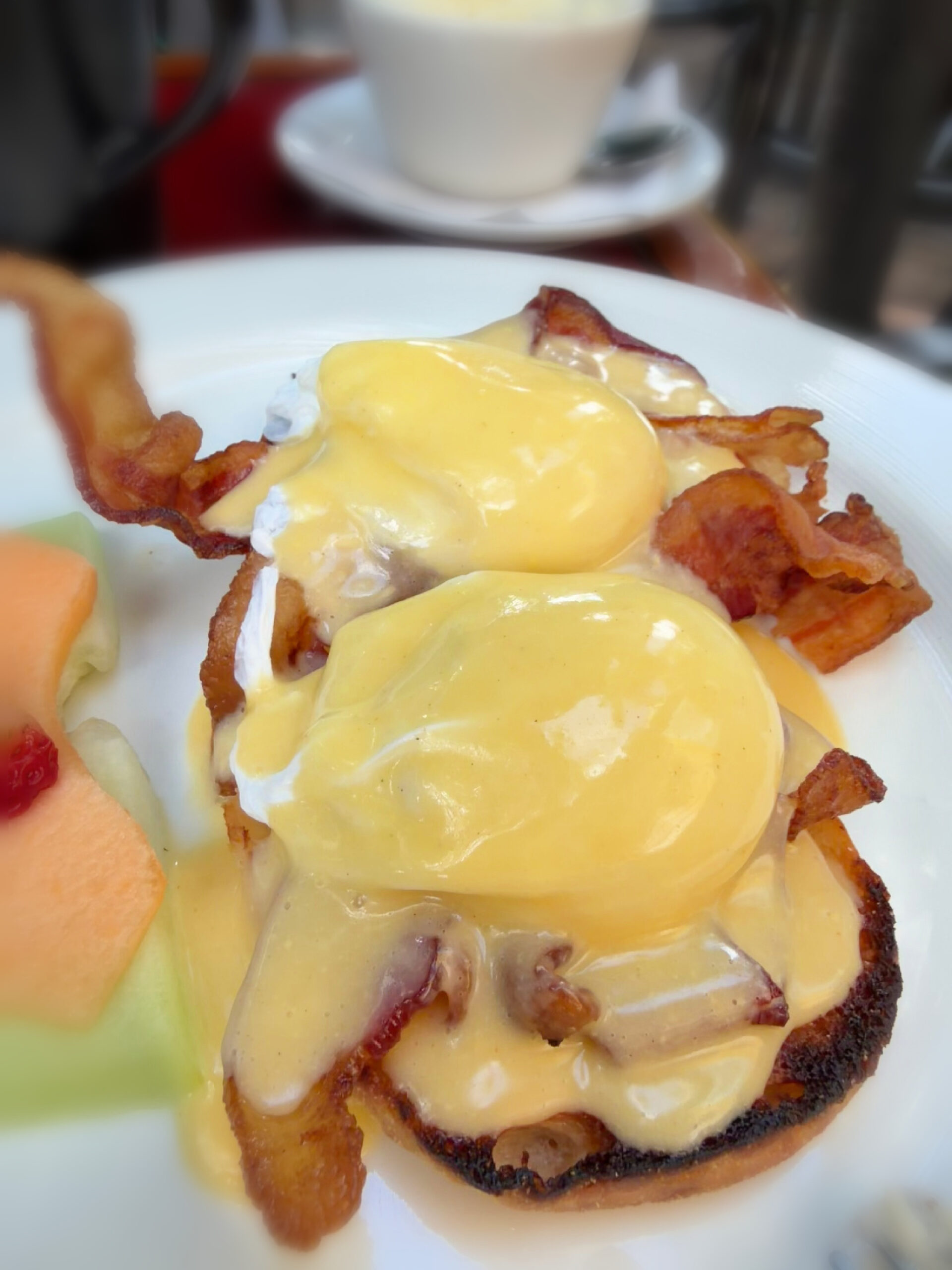 Two eggs Benedict on toasted English muffins with crispy bacon and creamy hollandaise sauce, served on a white plate with fresh cantaloupe slices on the side—a delicious breakfast option for anyone searching where to eat in Key West.