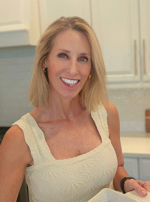 A woman with shoulder-length blonde hair, wearing a sleeveless beige textured dress, smiles while standing in a bright kitchen with white cabinets.