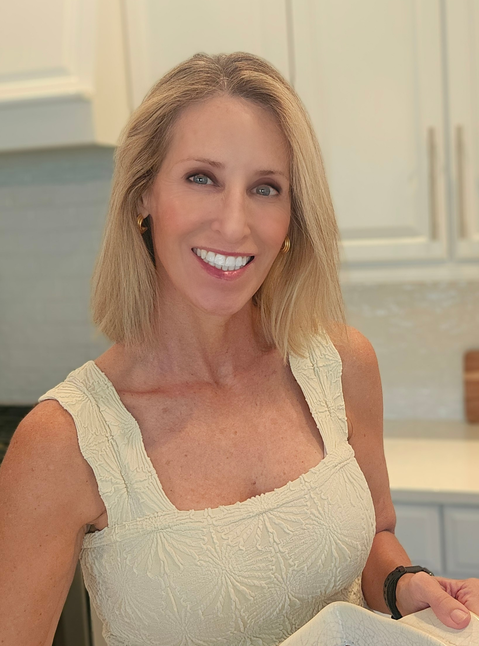 A woman with shoulder-length blonde hair, wearing a sleeveless beige textured dress, smiles while standing in a bright kitchen with white cabinets.