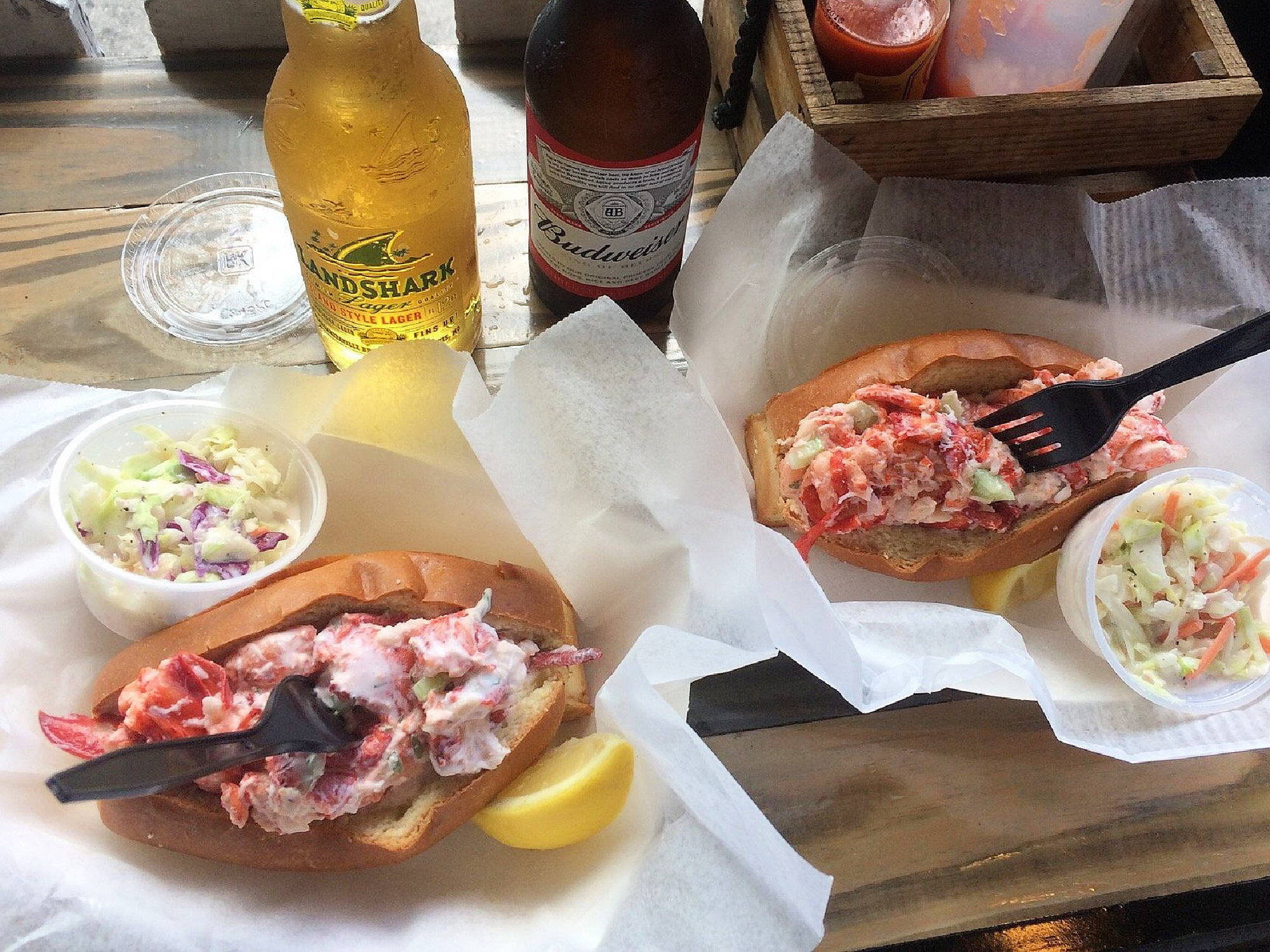 Savor two mouthwatering lobster rolls with coleslaw in small containers, lemon wedges, a chilled yellow LandShark beer, a Budweiser bottle, plastic forks, and condiments served on a rustic wooden table—a classic taste of where to eat in Key West.