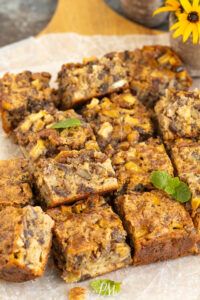 Nine golden-brown squares of apple and nut cake, reminiscent of classic apple date nut bread, are arranged on parchment paper. The cake pieces show a moist, chunky texture with visible apple and nut pieces. A small mint sprig and yellow flower decorate the scene.