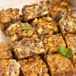 Nine golden-brown squares of apple and nut cake, reminiscent of classic apple date nut bread, are arranged on parchment paper. The cake pieces show a moist, chunky texture with visible apple and nut pieces. A small mint sprig and yellow flower decorate the scene.
