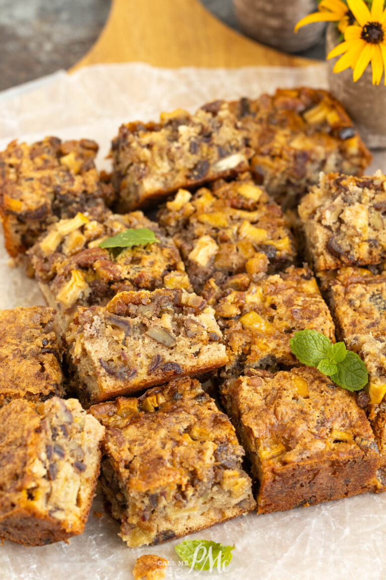 Nine golden-brown squares of apple and nut cake, reminiscent of classic apple date nut bread, are arranged on parchment paper. The cake pieces show a moist, chunky texture with visible apple and nut pieces. A small mint sprig and yellow flower decorate the scene.