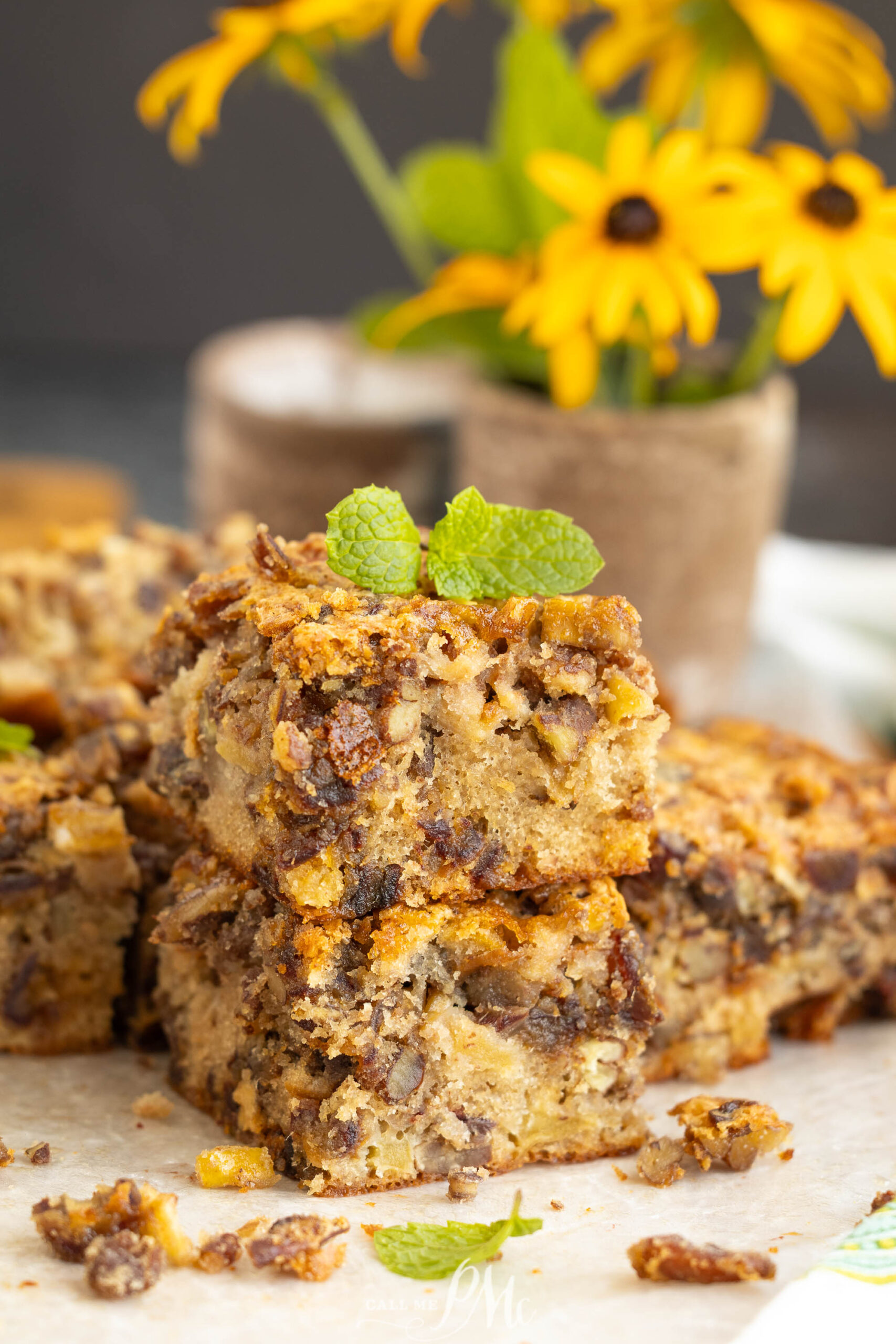 Two stacked blondie dessert bars. Yellow flowers in a vase are blurred in the background.