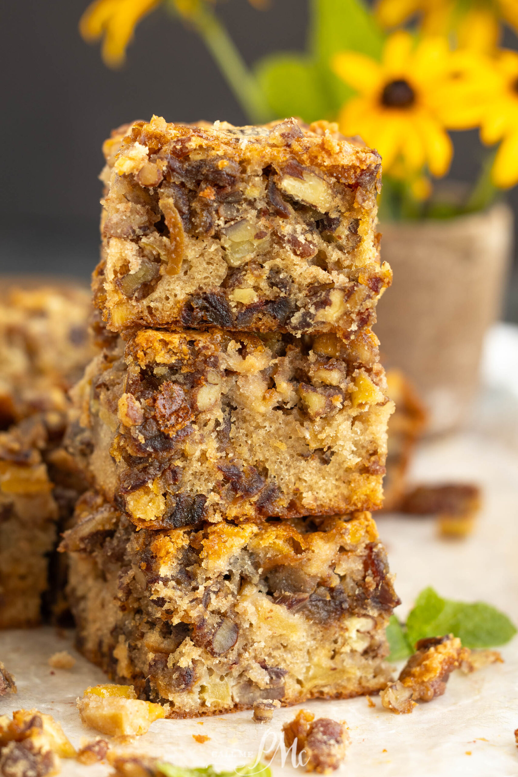 Three stacked squares of chewy bars with visible nuts and dried fruit, reminiscent of apple date nut bread, set on parchment paper. A blurred bouquet of yellow flowers is in the background.