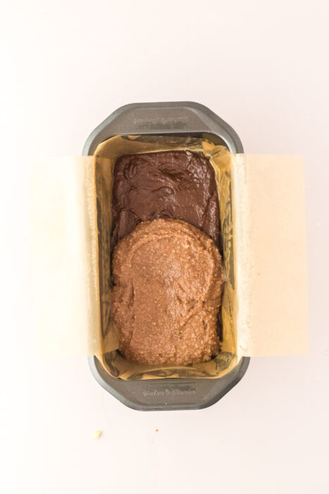 A loaf pan lined with parchment paper holds two types of batter, one brown and one light brown, side by side.