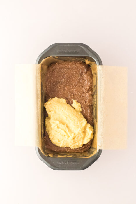 A loaf pan lined with parchment paper contains chocolate and yellow batter, partially mixed but mostly separated.
