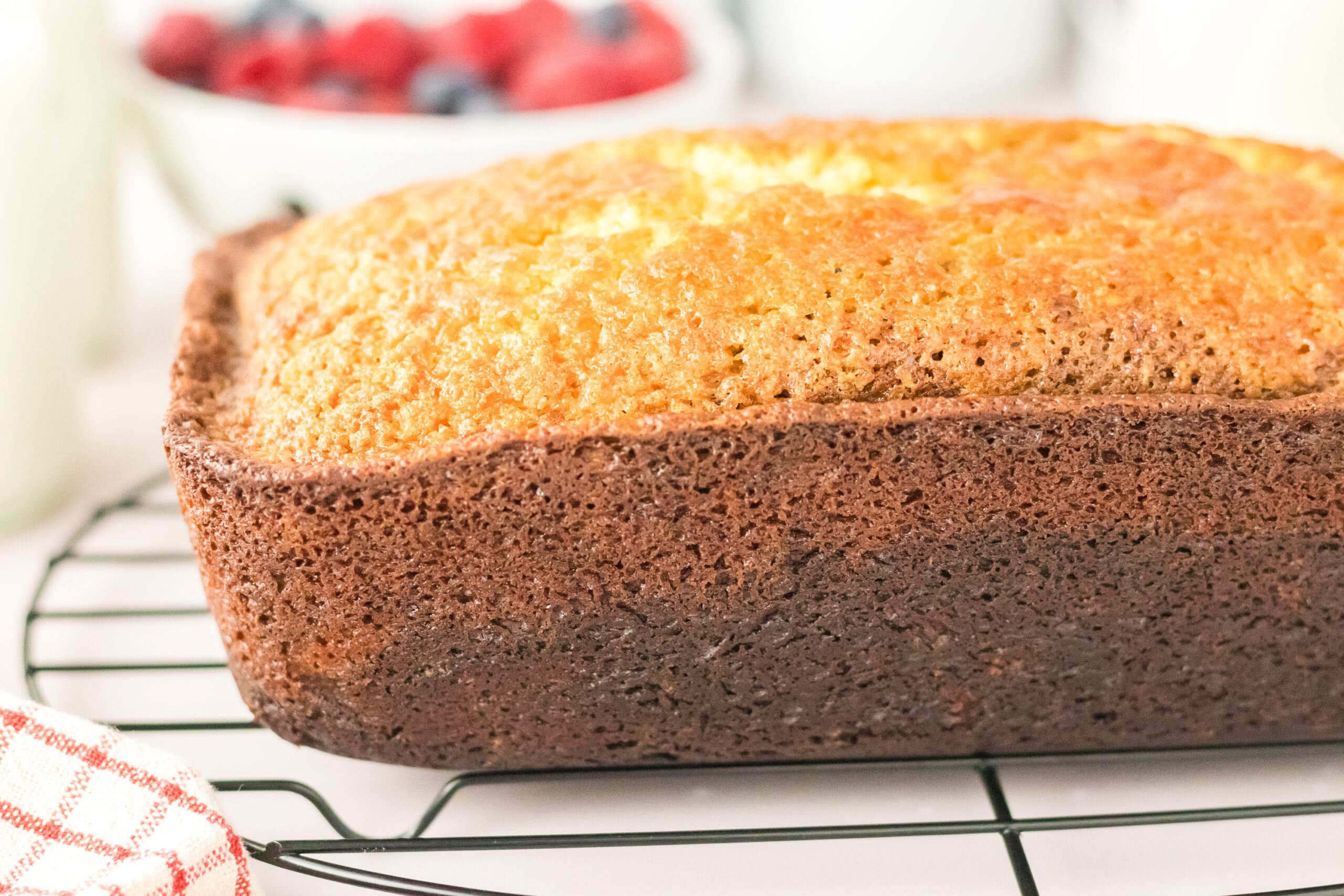 A loaf of golden brown Chocolate Ombre Pound Cake cooling on a wire rack, with a blurred bowl of mixed berries in the background.