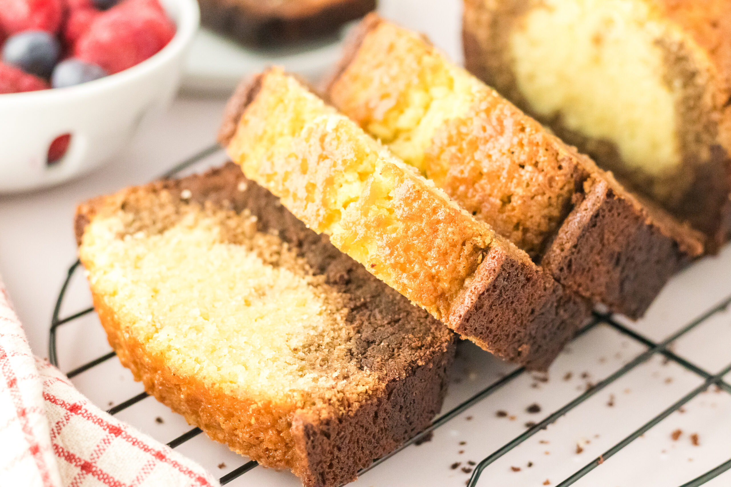 Three slices of Chocolate Ombre Pound Cake rest on a cooling rack, displaying a beautiful mix of chocolate and vanilla layers. A bowl of berries and a red-and-white towel are blurred in the background.