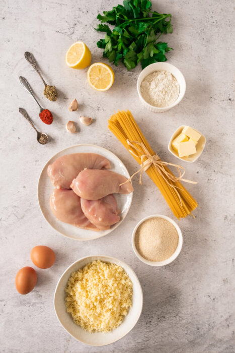 Raw chicken breasts, uncooked pasta, eggs, butter, flour, breadcrumbs, grated cheese, garlic cloves, herbs, lemon halves, and seasoning are arranged neatly on a light countertop—perfect for preparing crispy lemon garlic chicken.