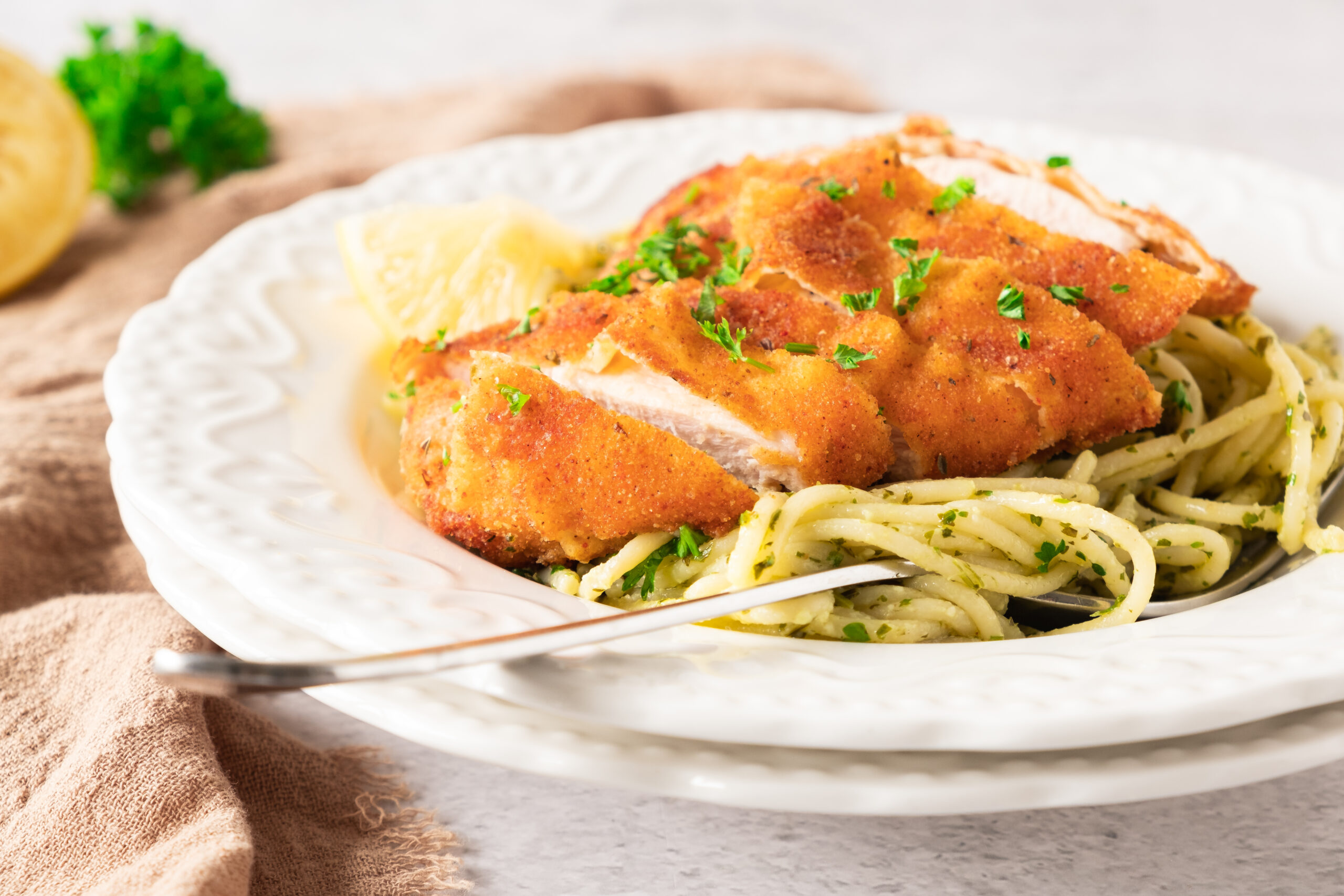 A plate of crispy lemon garlic chicken cutlets served on a bed of spaghetti tossed with pesto sauce, garnished with chopped parsley and a lemon wedge. A fork rests on the side of the bowl.