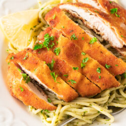 Crispy lemon garlic chicken cutlet sliced on top of spaghetti with green pesto sauce, garnished with chopped parsley and a lemon wedge on a white plate.