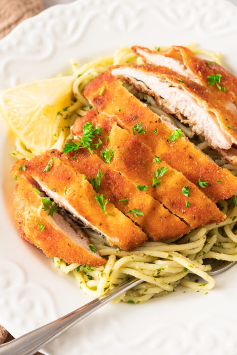 Crispy lemon garlic chicken cutlet sliced on top of spaghetti with green pesto sauce, garnished with chopped parsley and a lemon wedge on a white plate.