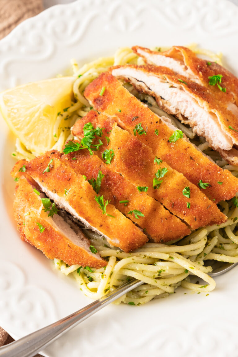 Crispy lemon garlic chicken cutlet sliced on top of spaghetti with green pesto sauce, garnished with chopped parsley and a lemon wedge on a white plate.