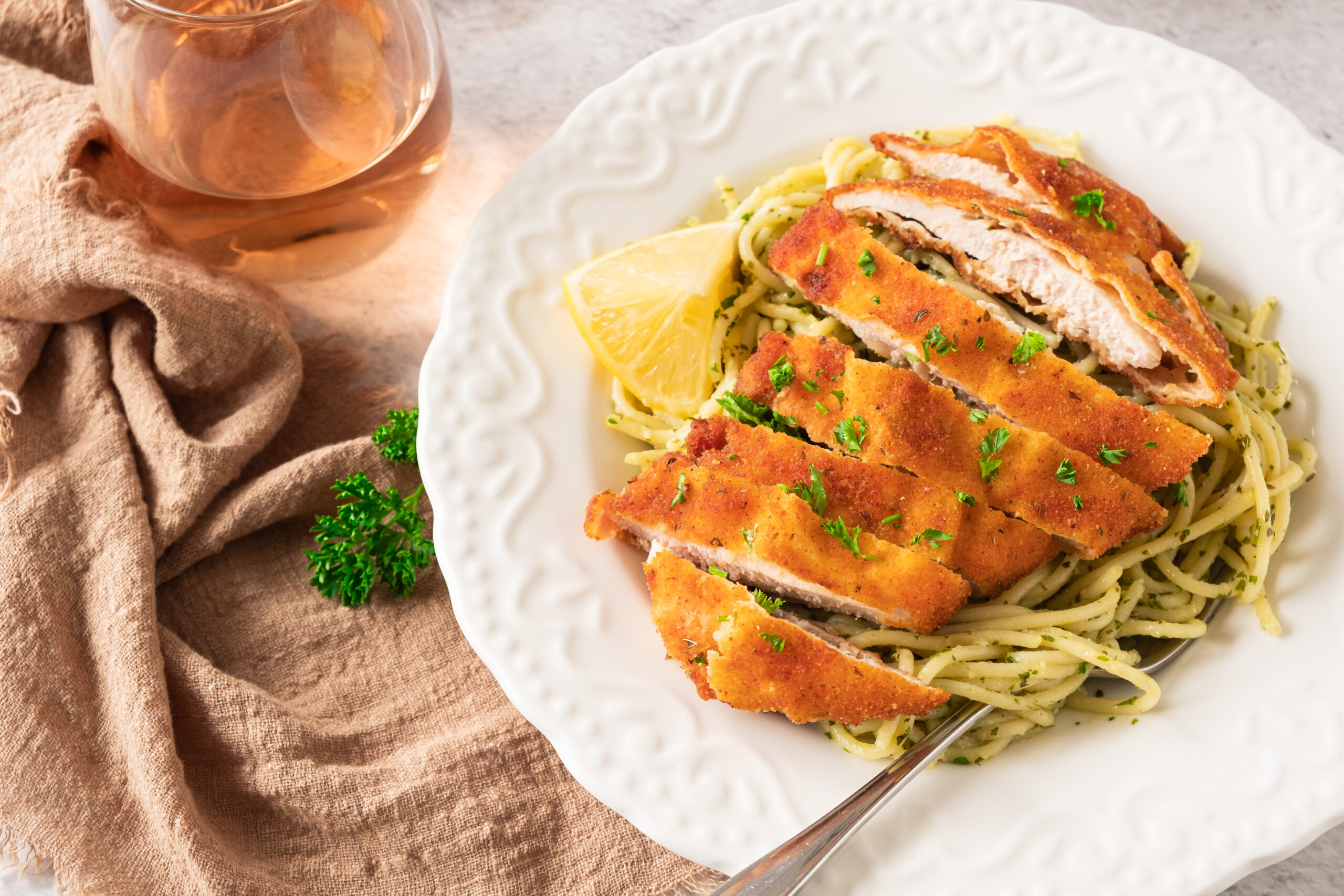 A plate of crispy lemon garlic chicken cutlets served on a bed of spaghetti tossed with pesto sauce, garnished with chopped parsley and a lemon wedge. A fork rests on the side of the bowl.