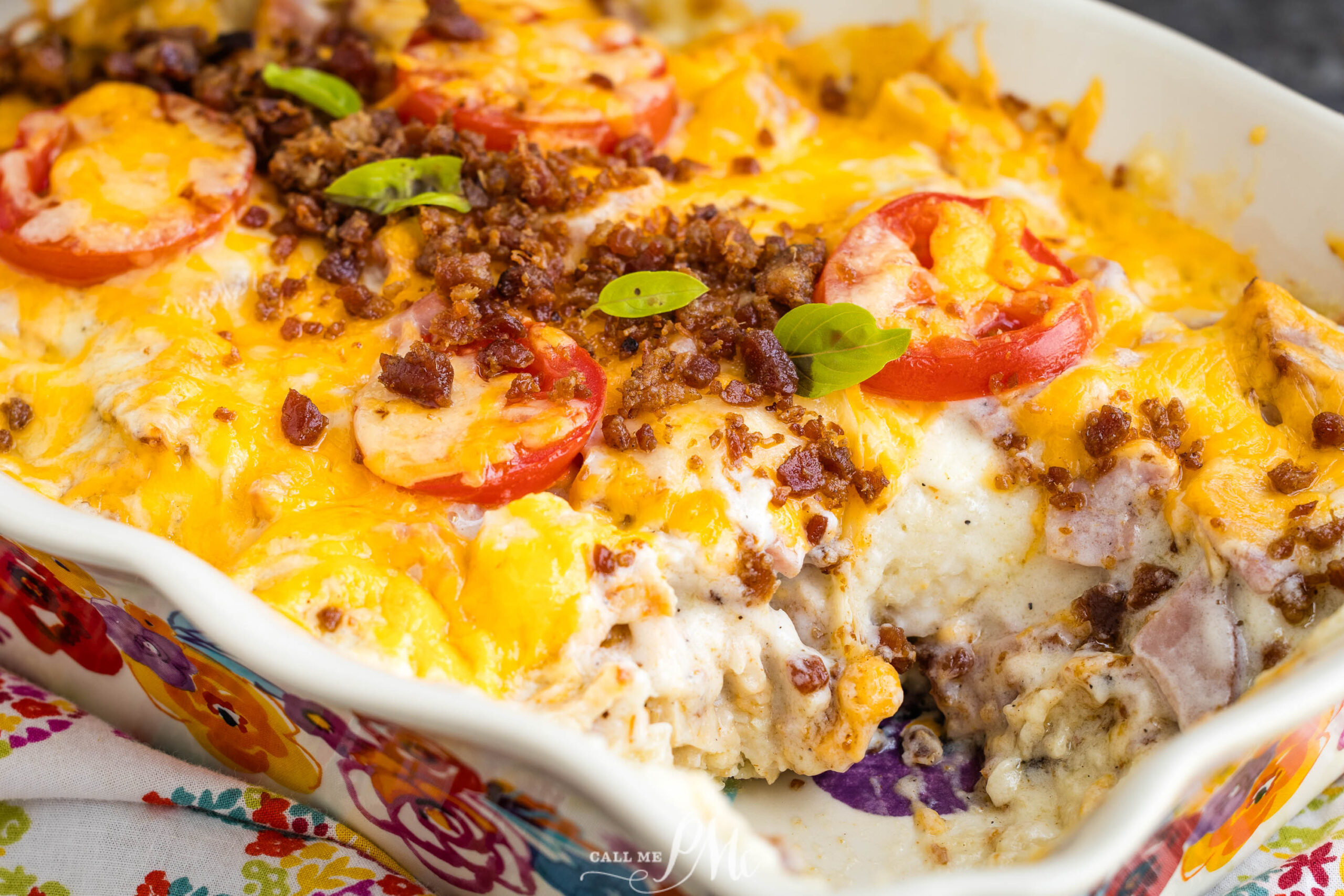 A baked casserole topped with melted cheddar cheese, sliced tomatoes, bacon bits, and fresh green herbs, with a portion already served, revealing creamy layers inside.