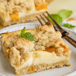 A square of Peach Cheesecake Crumb Bar with a creamy filling and crumbly topping sits on a plate, garnished with a mint leaf. A fork rests beside it, and another dessert piece is visible in the background.