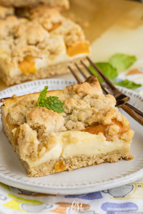 A square of Peach Cheesecake Crumb Bar with a creamy filling and crumbly topping sits on a plate, garnished with a mint leaf. A fork rests beside it, and another dessert piece is visible in the background.