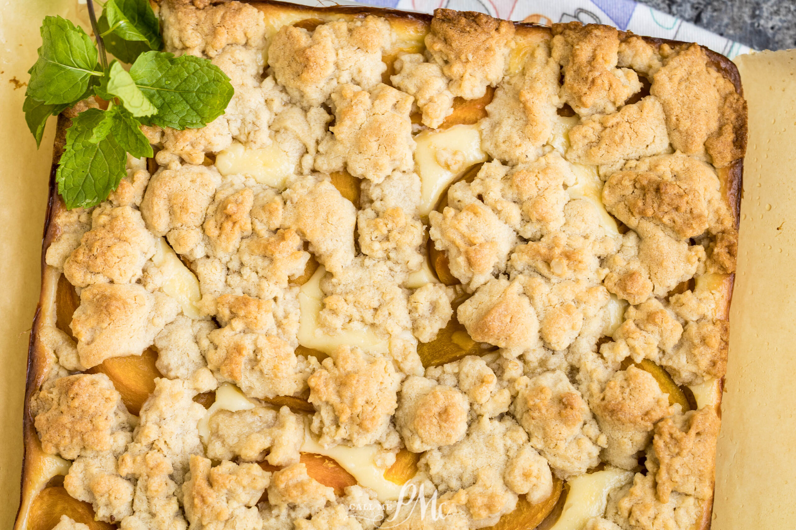 A close-up of Peach Cheesecake Crumb Bars on parchment paper, topped with golden, crumbly streusel and garnished with a sprig of fresh mint in the corner.