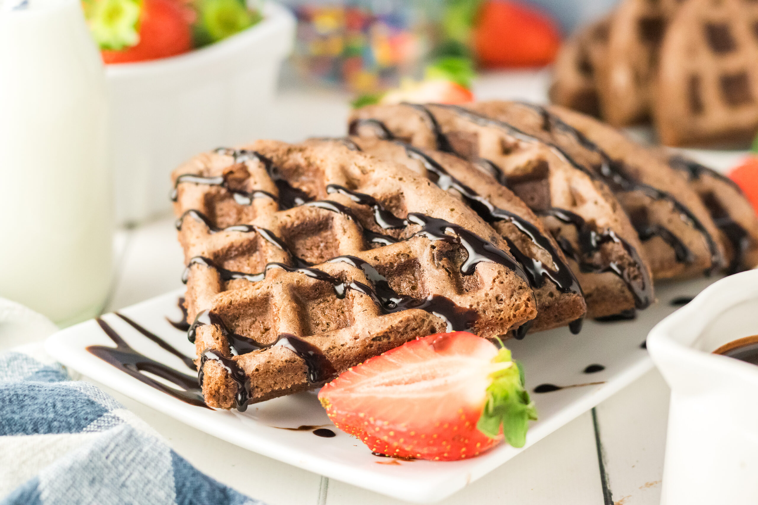 Brownie Mix Waffles drizzled with chocolate syrup are served on a white plate with a fresh strawberry. A glass of milk and more strawberries are in the background.