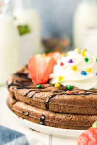 Close-up of Brownie Mix Waffles topped with whipped cream, rainbow sprinkles, chocolate syrup, and a sliced strawberry on a white plate. Milk bottles and fruit are blurred in the background.