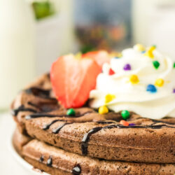 Close-up of Brownie Mix Waffles topped with whipped cream, rainbow sprinkles, chocolate syrup, and a sliced strawberry on a white plate. Milk bottles and fruit are blurred in the background.