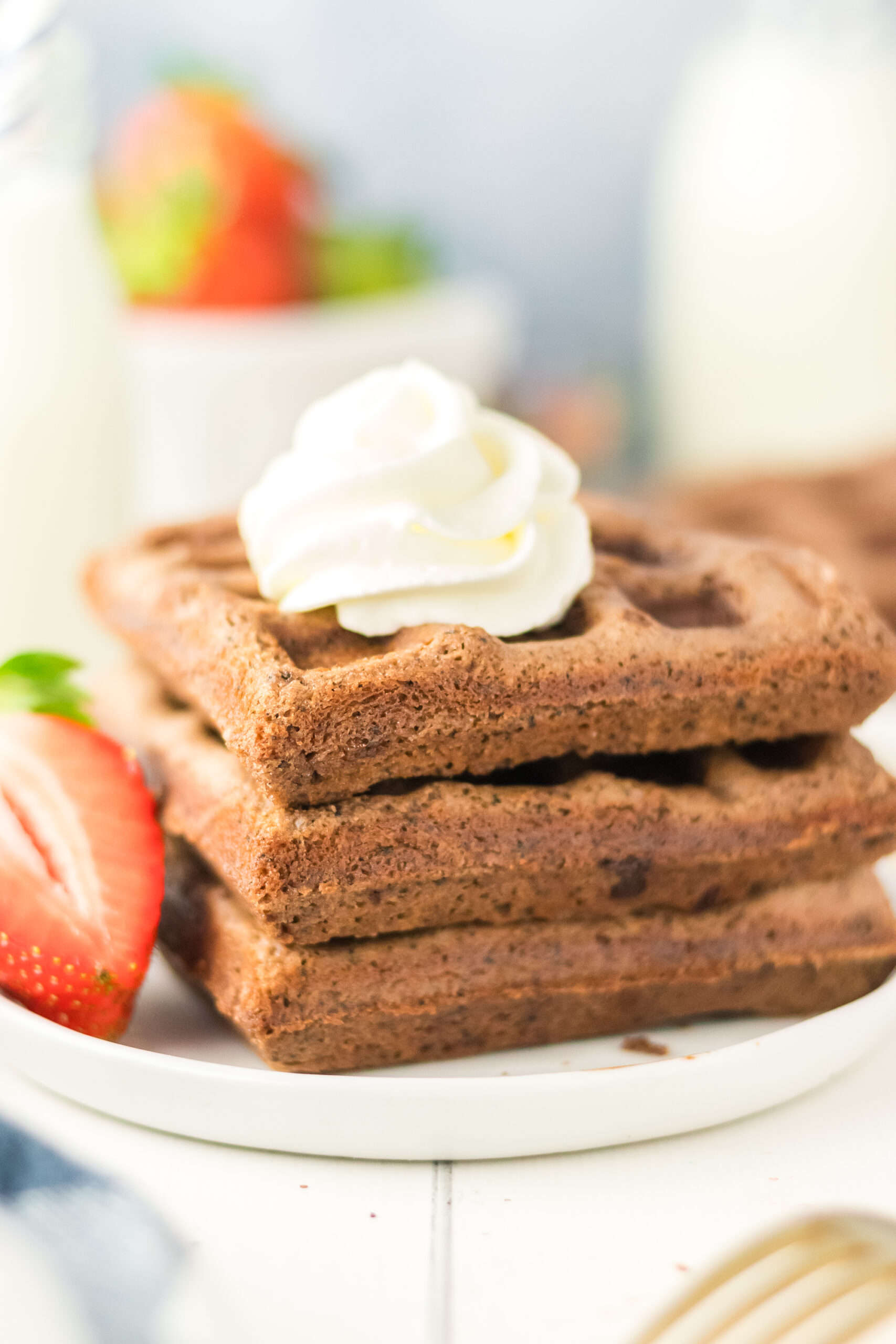Flapjacks stacked on a white plate and topped with a dollop of whipped cream. A sliced strawberry rests on the side, with blurred milk bottles and fresh strawberries in the background.