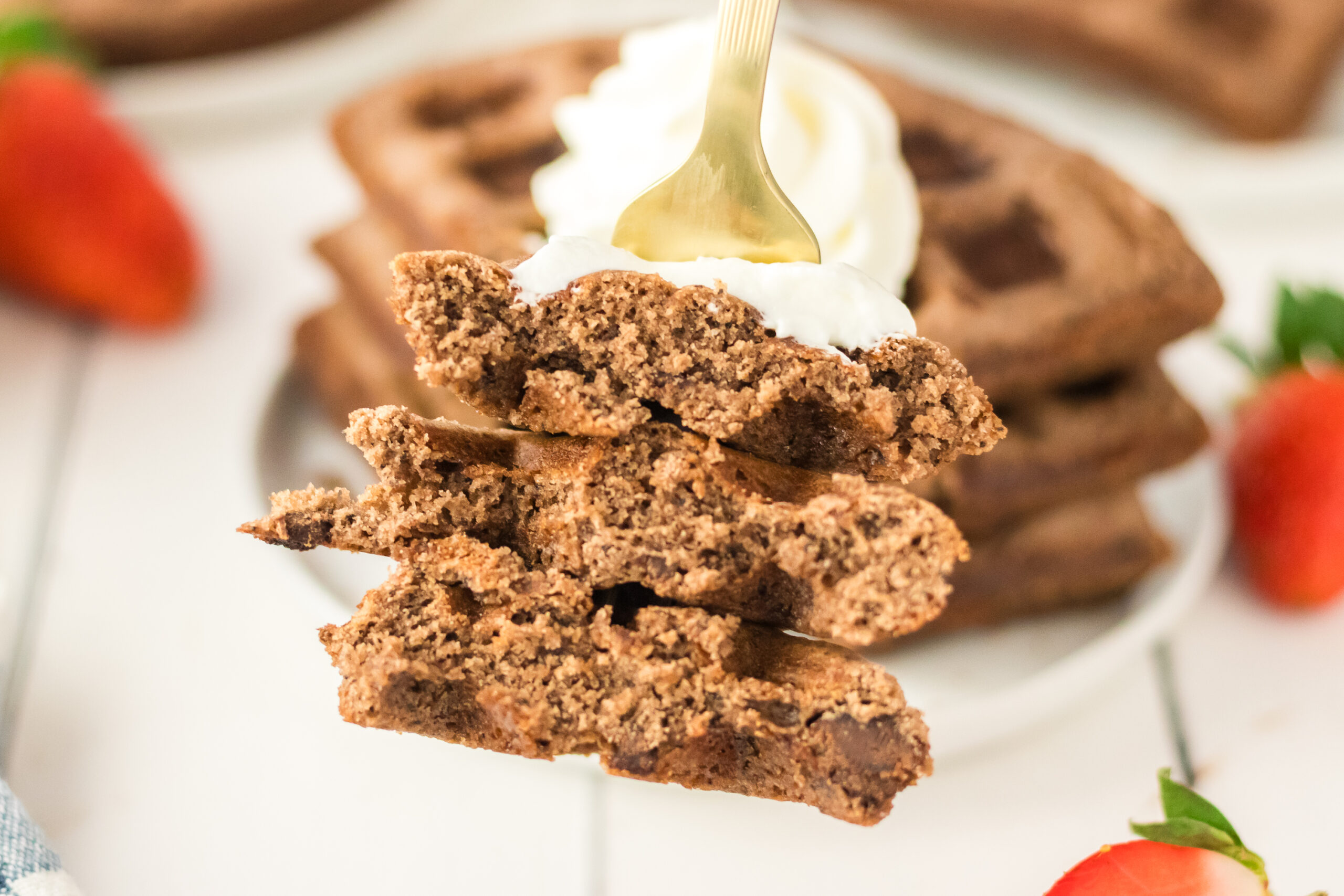 A fork holds pieces of chocolate waffle topped with whipped cream, made even more decadent by using Brownie Mix Waffles. More waffles and strawberries are blurred in the background.