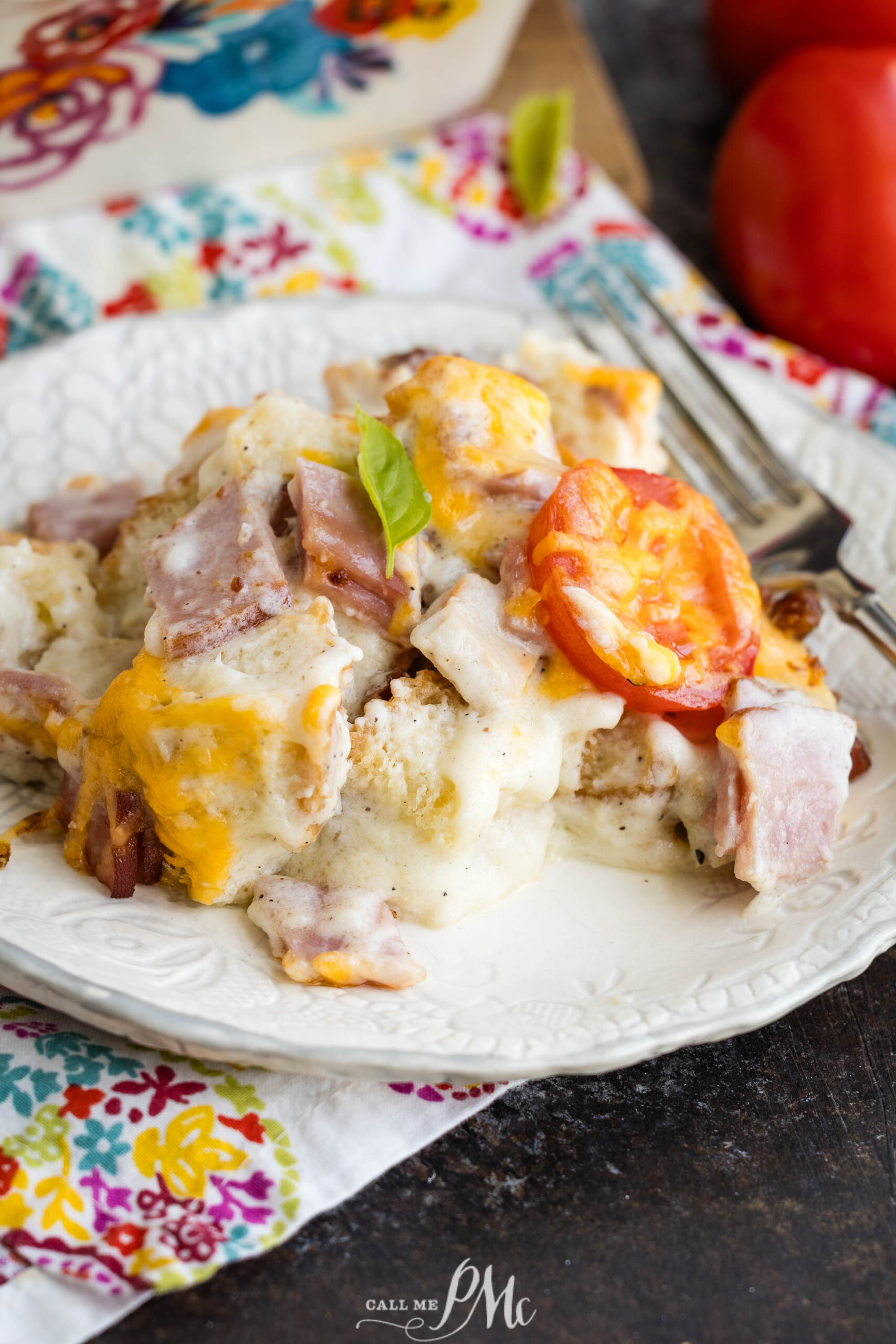 A serving of cheesy ham and tomato casserole, reminiscent of a classic Kentucky Hot Brown Casserole, sits on a white plate, garnished with a basil leaf next to a fork. The background includes fresh tomatoes and a colorful floral napkin.
