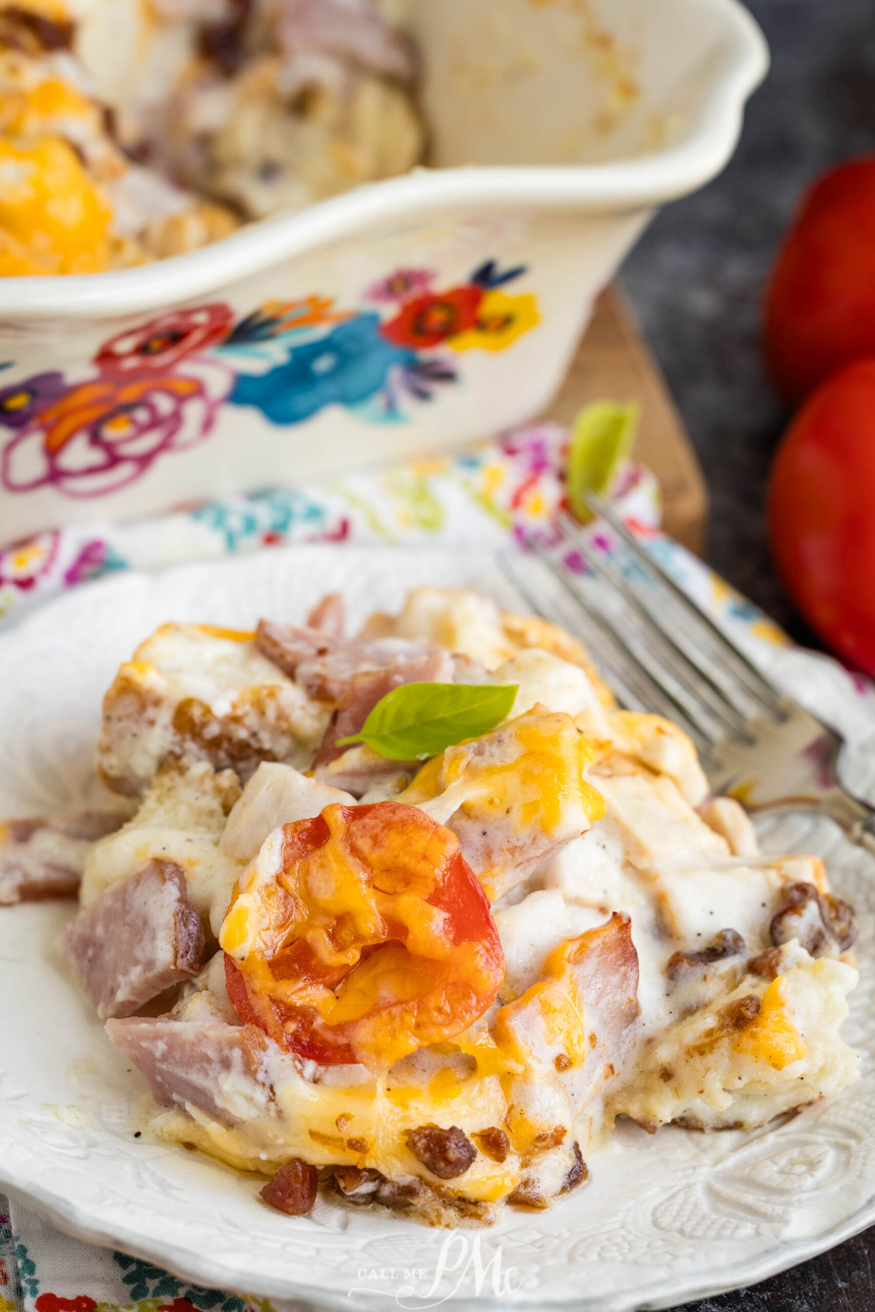 A serving of cheesy breakfast casserole with ham, tomato, and herbs sits on a white plate. Reminiscent of a Kentucky Hot Brown Casserole, this dish is hearty and comforting. A fork rests nearby, and the floral casserole dish is partially visible in the background.