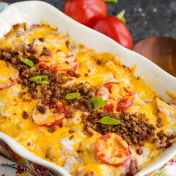 A baked casserole topped with melted cheese, sliced tomatoes, ground meat, and green herbs, served in a decorative dish with fresh tomatoes and green onions in the background.