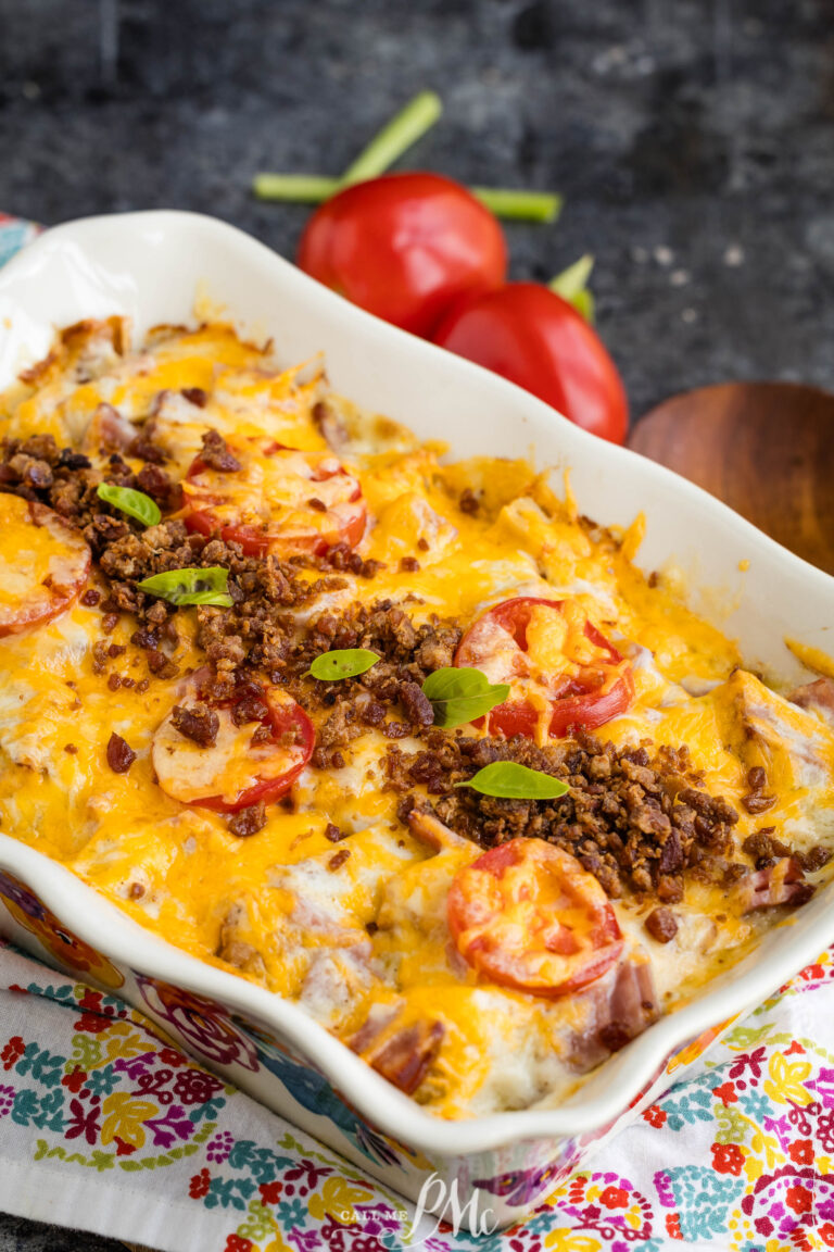 A baked casserole topped with melted cheese, sliced tomatoes, ground meat, and green herbs, served in a decorative dish with fresh tomatoes and green onions in the background.