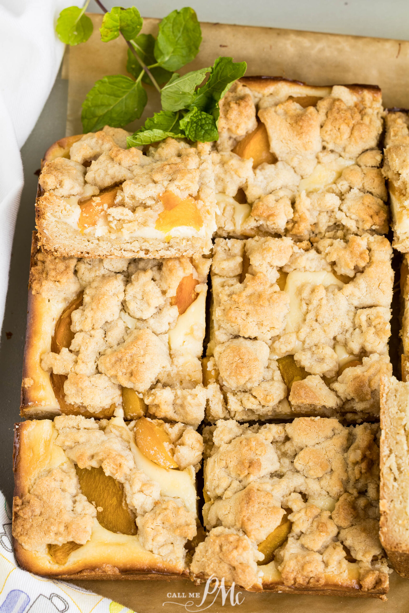 Peach Cheesecake Crumb Bars cut into squares rest on parchment paper, showcasing visible peach slices layered beneath a crumbly topping. A sprig of mint is placed beside the bars for garnish, adding a fresh touch to these delightful treats.