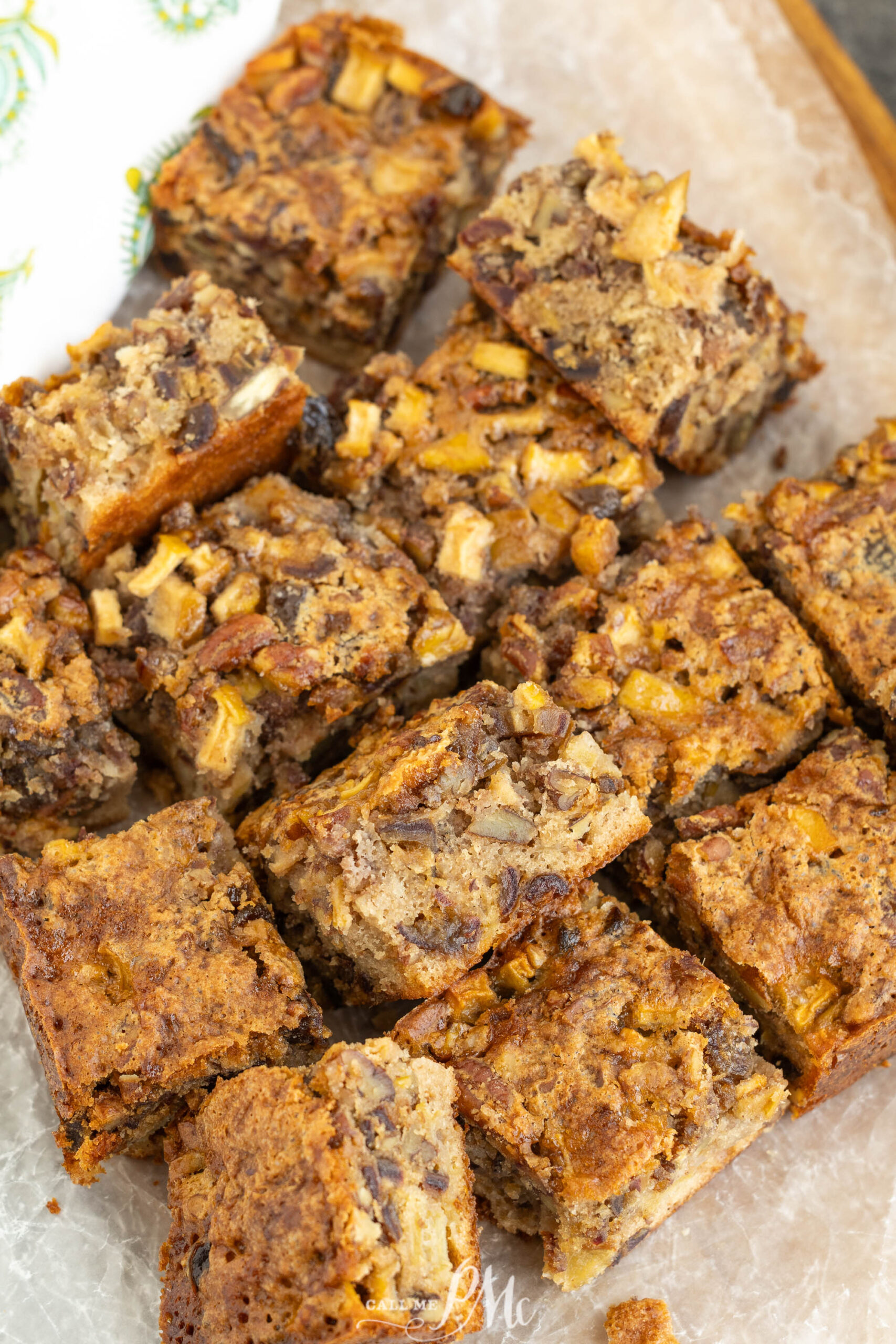 Squares of apple pecan cake are arranged on parchment paper, showing a moist texture with visible chunks of apples and pecans throughout the golden-brown cake—reminiscent of a classic apple date nut bread, but with a delightful twist.