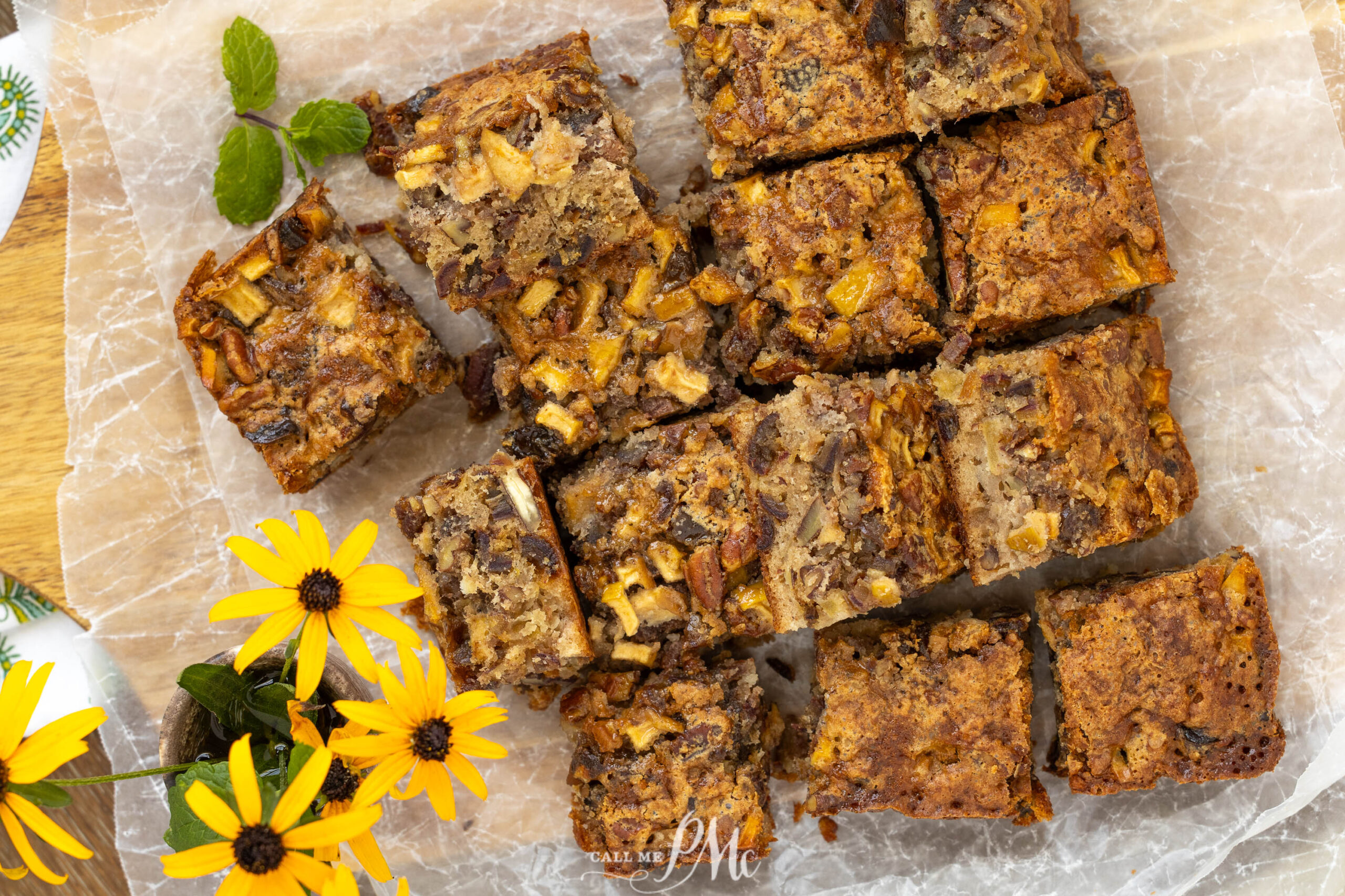 Sliced apple pecan cake bars arranged on parchment paper, reminiscent of classic apple date nut bread, and garnished with yellow flowers and green leaves.