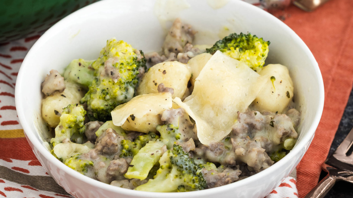 A white bowl filled with gnocchi, broccoli, ground meat, and a creamy sauce, topped with shaved cheese. The bowl sits on a patterned cloth.