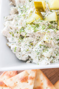 Close-up of creamy dill pickle chicken salad with herbs and diced pickles in a white dish, with crackers visible in the background.