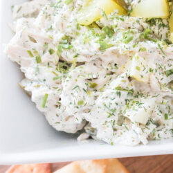 Close-up of creamy dill pickle chicken salad with herbs and diced pickles in a white dish, with crackers visible in the background.