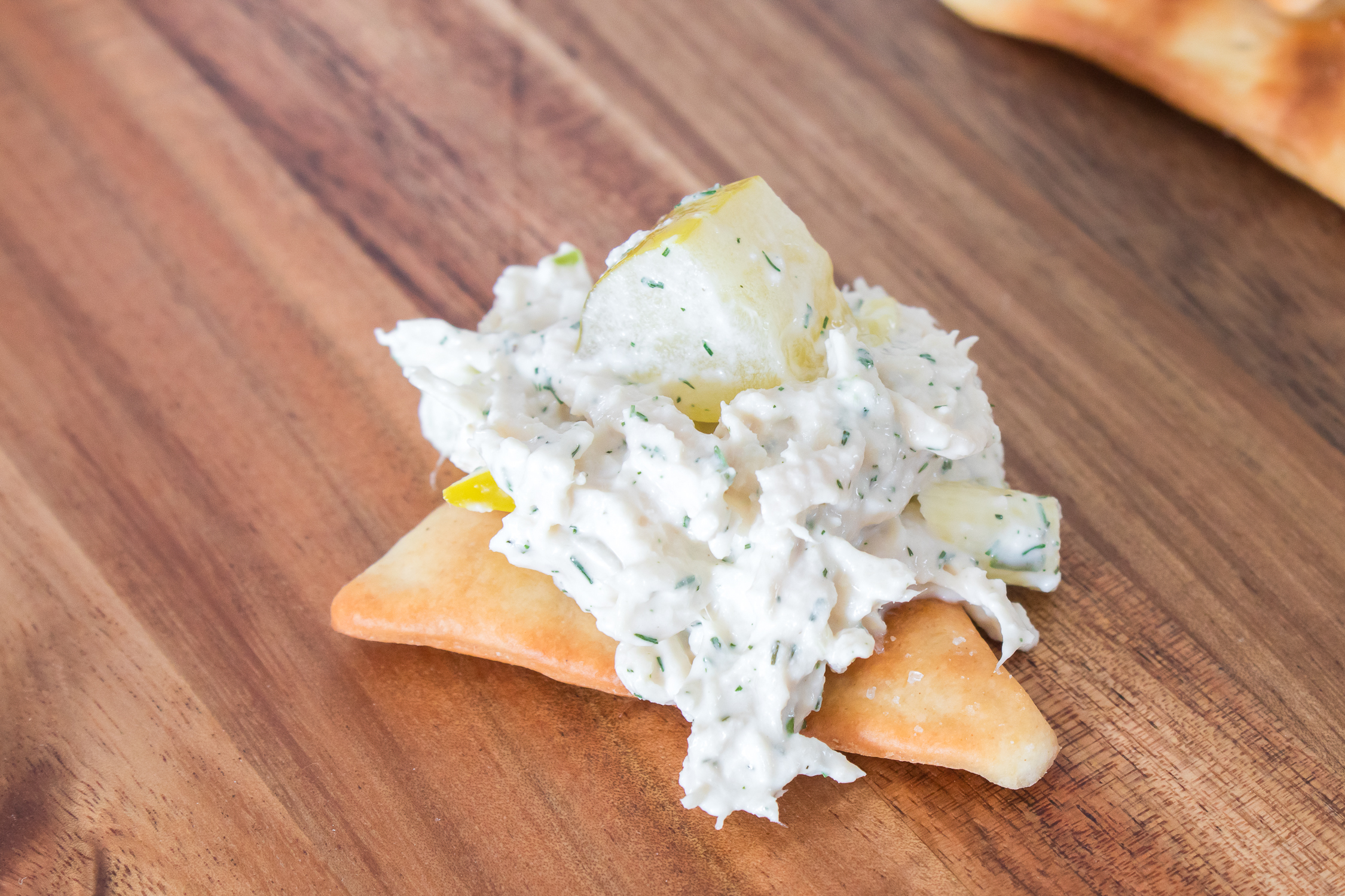 A square cracker topped with a creamy dill pickle chicken salad mixed with herbs and a chunk of yellow pickle, placed on a wooden surface.