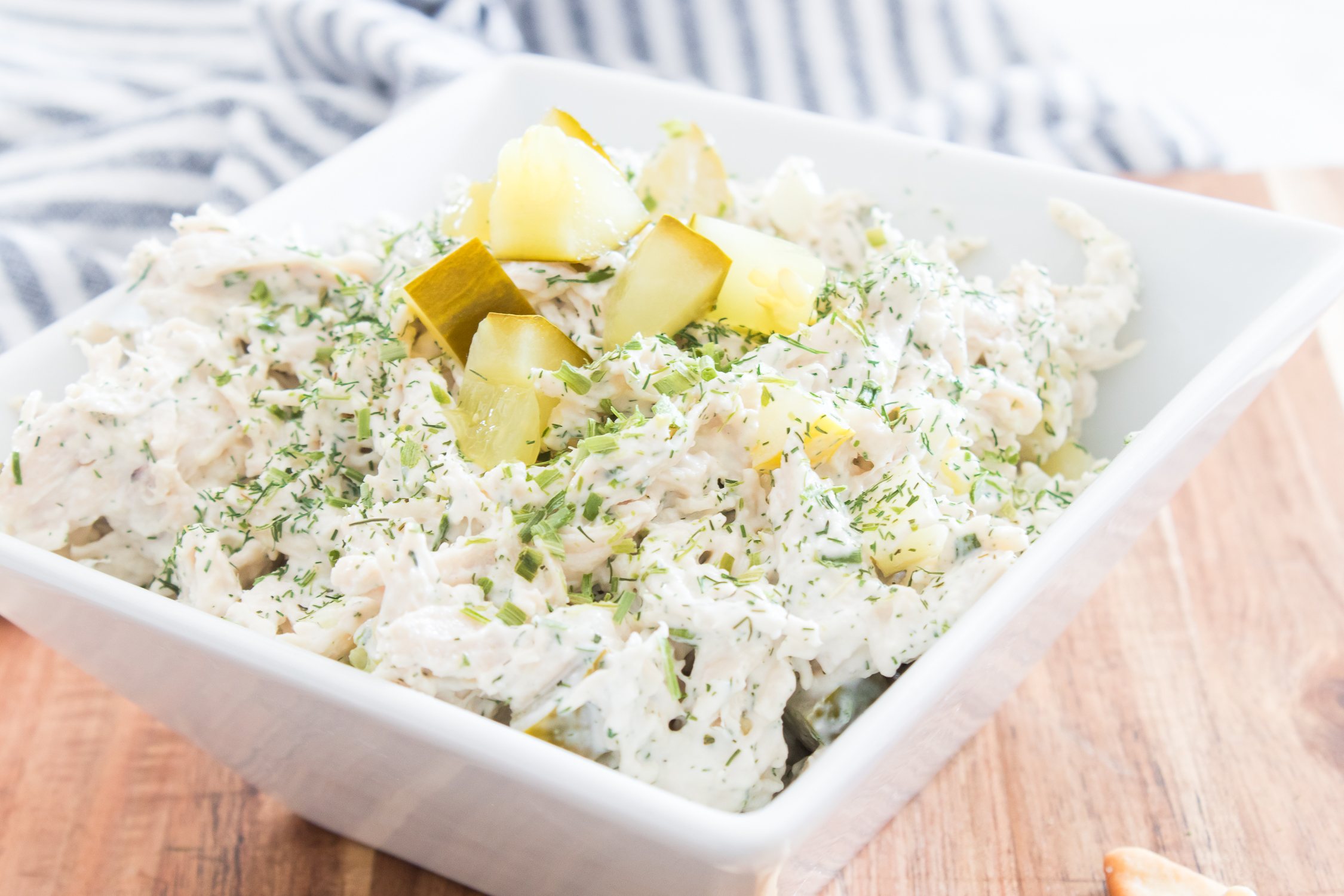 Dill pickle chicken salad with chopped pickles and herbs, served in a white square bowl on a wooden surface. A striped cloth is visible in the background.