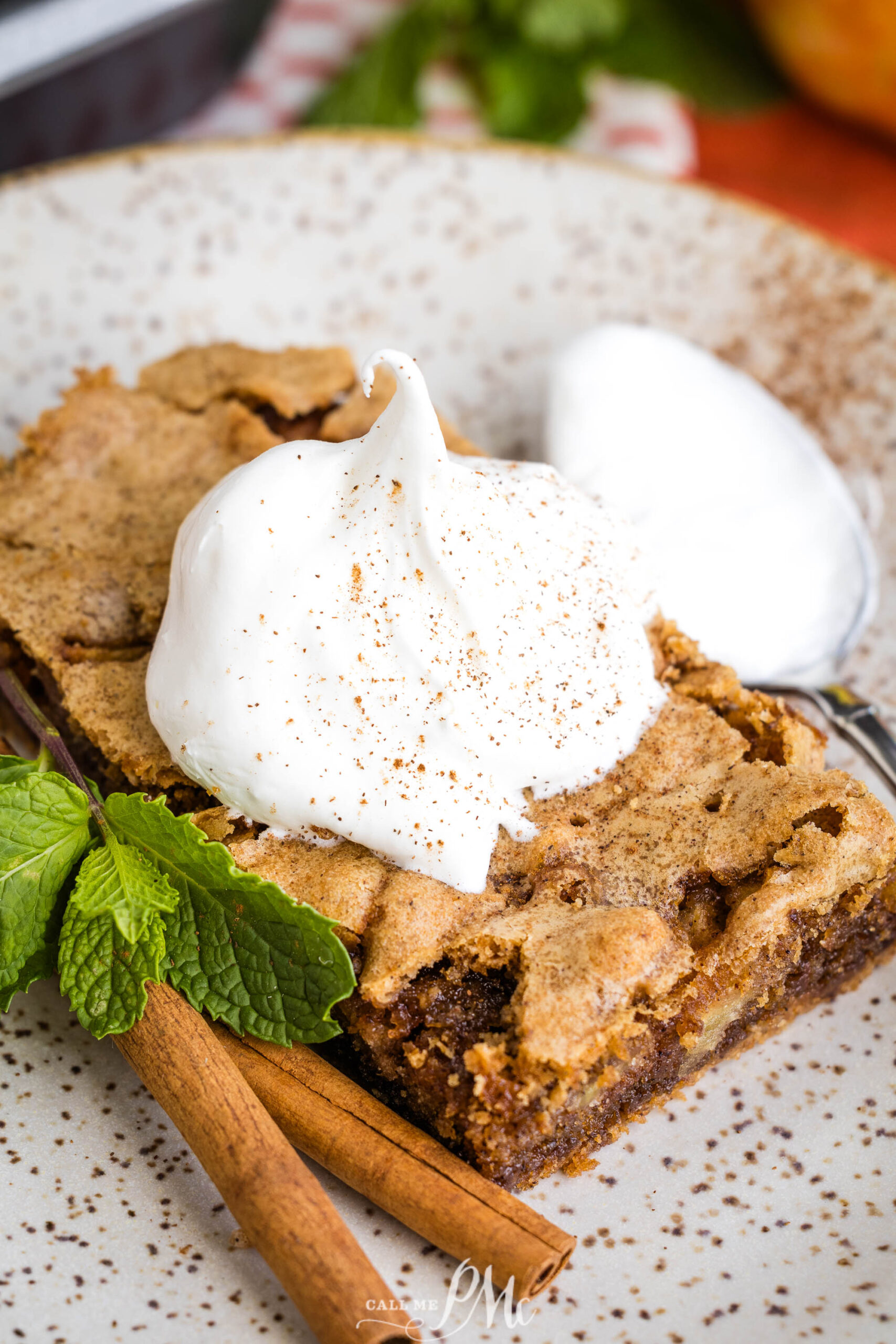 A slice of dessert topped with whipped cream and sprinkled spices, garnished with fresh mint and cinnamon sticks, served on a speckled plate with a spoon in the background.