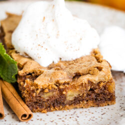 A square slice of German Apple Cake topped with a dollop of whipped cream and a sprinkle of cinnamon, served on a plate with a cinnamon stick and a mint leaf.
