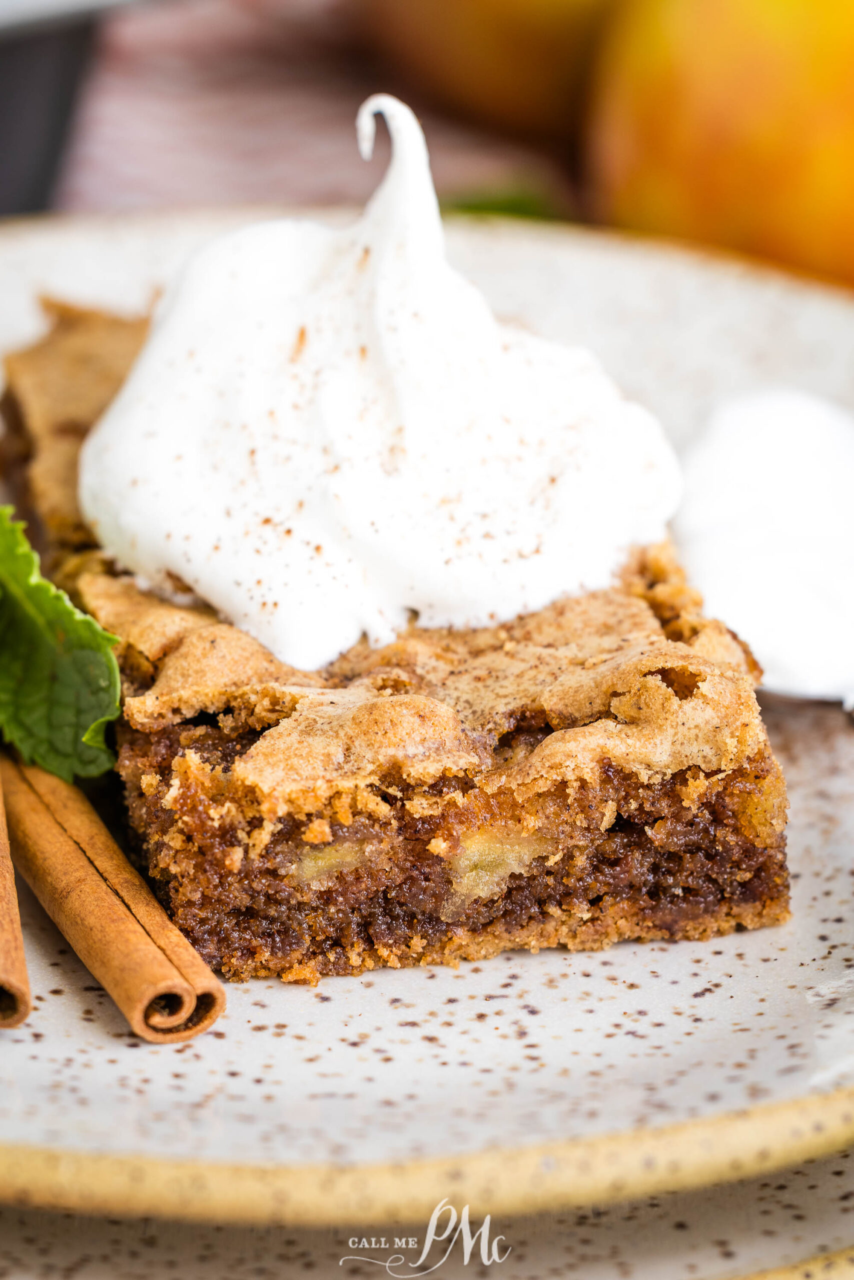 A square slice of dessert topped with a dollop of whipped cream and a sprinkle of cinnamon, served on a plate with a cinnamon stick and a mint leaf.