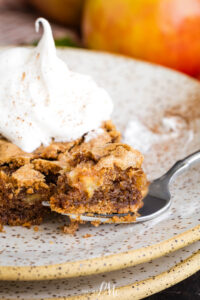 A fork with a bite of spiced cake rests on a speckled plate, topped with a swirl of whipped cream. In the background, a blurred apple hints at the classic flavors of German Apple Cake throughout the scene.