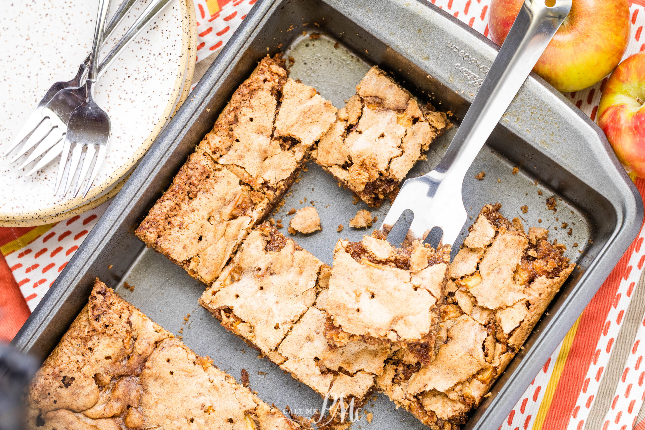 A baking pan with sliced, golden-brown German Apple Cake dessert bars on a table, surrounded by apples, a plate with forks, and a spatula lifting one bar from the pan.