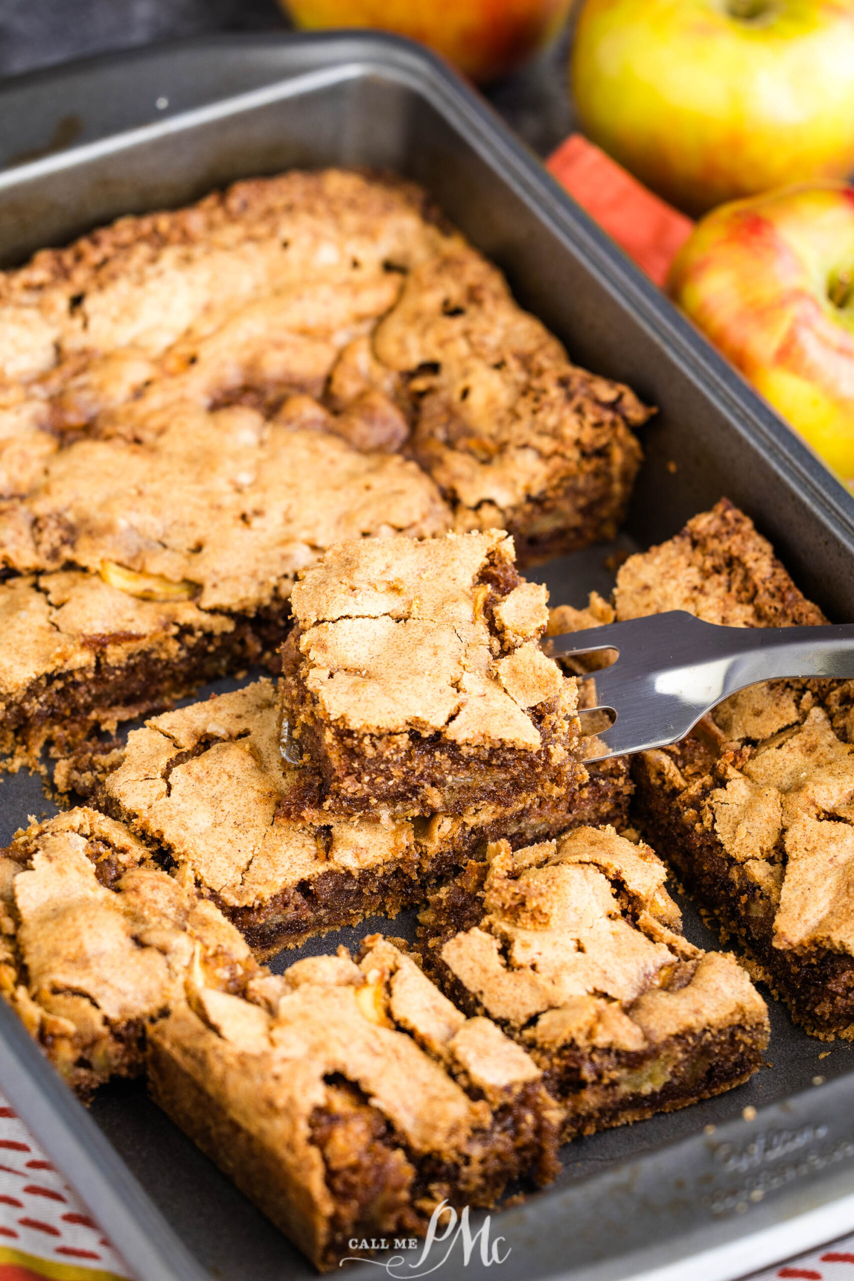 A fork lifts a square slice of golden-brown German Apple Cake blondie from a baking pan filled with similar pieces. Fresh apples are visible in the background.