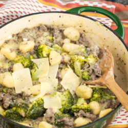 A green pot filled with gnocchi, broccoli, ground meat, and topped with shaved Parmesan cheese. A wooden spoon rests inside the pot, which sits on a red and white striped cloth.