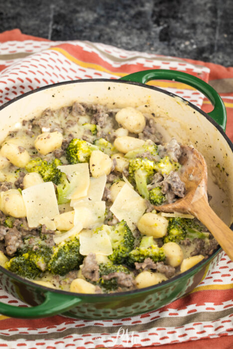 A green pot filled with gnocchi, broccoli, ground meat, and topped with shaved Parmesan cheese. A wooden spoon rests inside the pot, which sits on a red and white striped cloth.